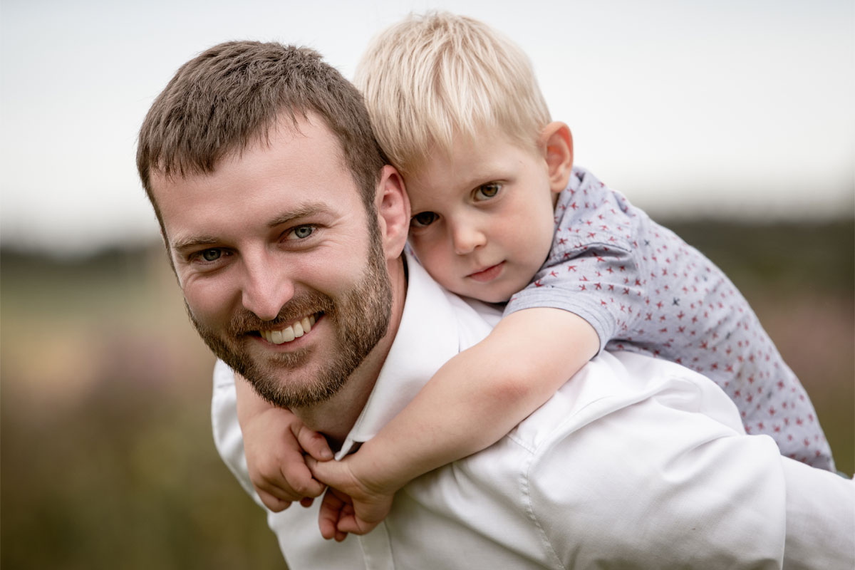 Vater trägt seinen jungen Sohn am Rücken, beide eng aneinander, der Vater lächelt herzlich in die Kamera, während der Bub ruhig und aufmerksam blickt.